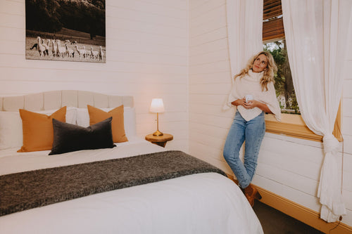 A woman is standing in a cottage bedroom, leaning against the windowsill, wearing an alpaca poncho.