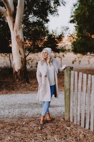 A woman stands next to a fence wearing a beige alpaca coat over a cream alpaca knit, styled with cream alpaca accessories, light denim jeans and boots.